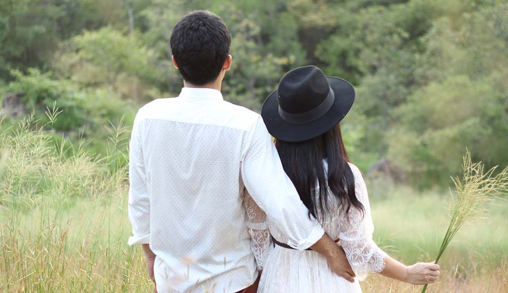 Pareja de espaldas mirando la montaña, él con la mano en la cintura de ella, ella sosteniendo una rama